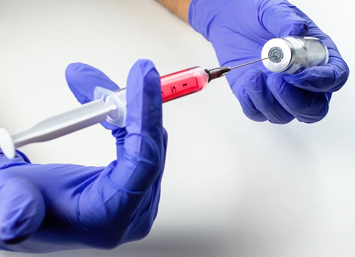 Hands in blue gloves are holding a disposable syringe and an ampoule with red liquid (Vitamin B12)
